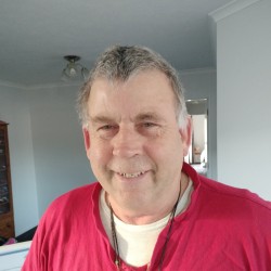 Photo of head and shoulders of an older man with short grey hair, smiling, wearing a red top with a white t-shirt underneath.